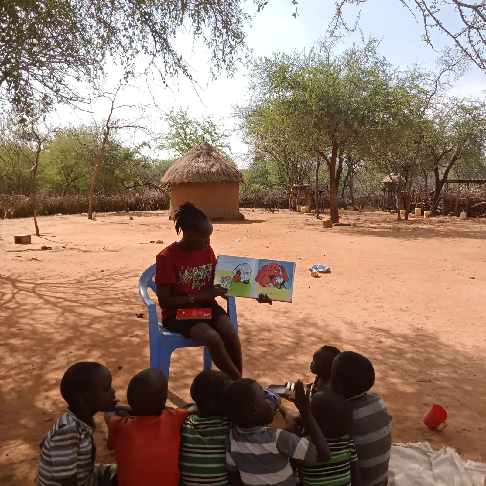 A teacher in a red top reads a picture book to children seated on the ground under trees beside a thatched hut