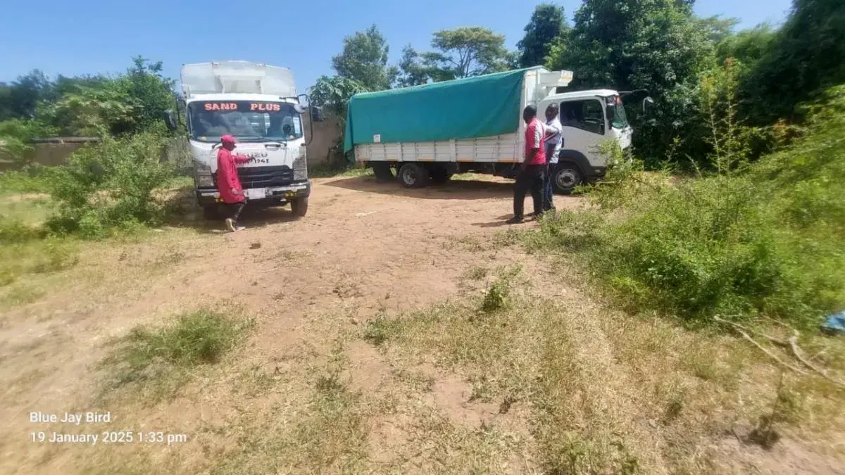 Two lorries parked on a dusty track beside green bushes, with a person in red standing by the nearer one