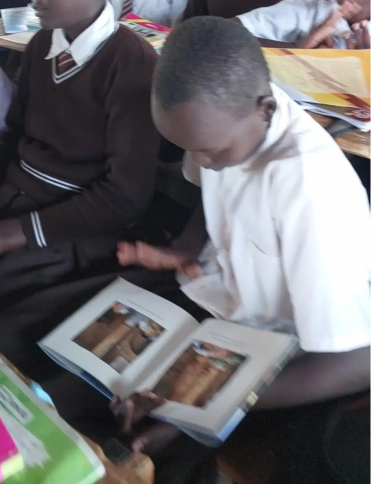 A pupil in a white shirt looks through a picture book on his lap, with other pupils in dark school jumpers beside him