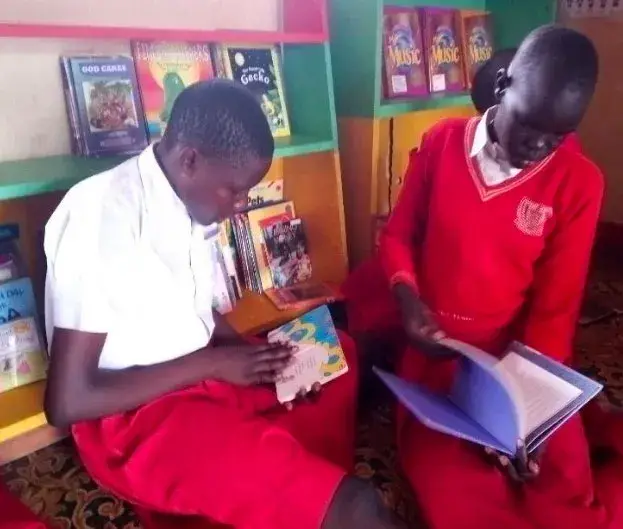 Two pupils in red school uniform sitting on the floor of a school library reading picture books, with shelves of books behind them