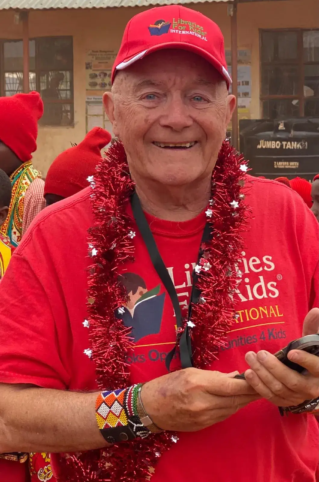 Roy Austin, the founder, smiling in a red Libraries For Kids cap and T-shirt with a red tinsel garland around his neck and a beaded bracelet on his wrist, in a school yard with children in red hats behind him