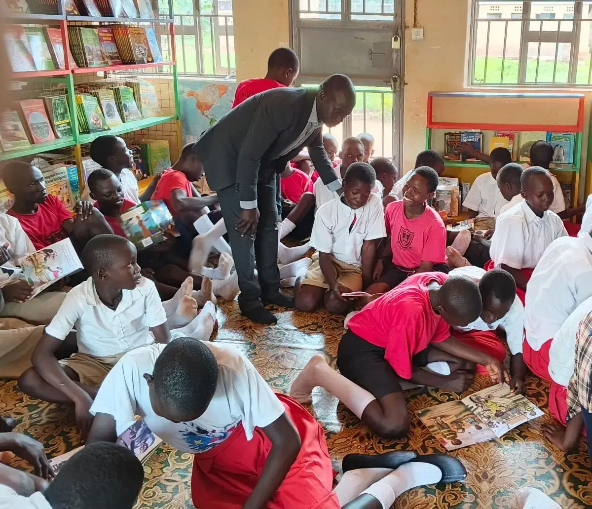Many pupils in red and white school uniforms sitting on the floor of a library reading books, with a man in a dark suit leaning over to help and shelves of books behind them