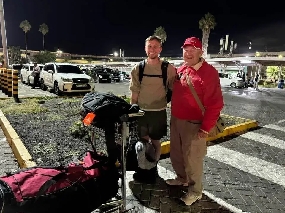 Two men standing in a night-time car park beside luggage: a younger man with a backpack, and an older man in a red cap and red shirt