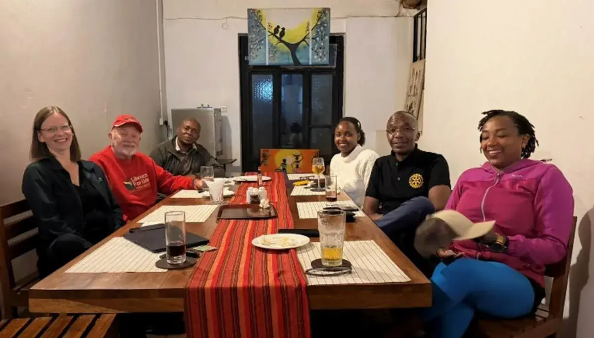 Seven people seated around a long table laid with a red runner in a restaurant, at a Rotary meeting in Karatu