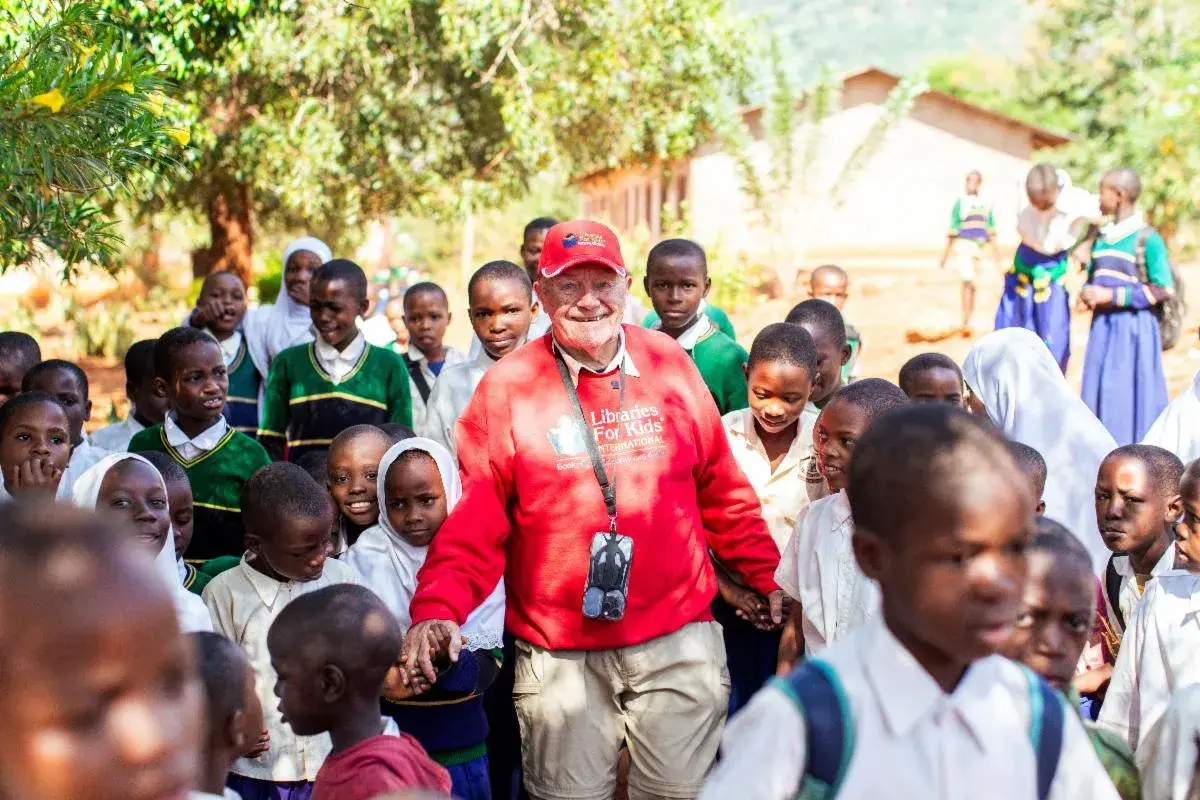 A man in a red jacket and cap, with binoculars on a strap, walking among a crowd of smiling schoolchildren in uniform outdoors, with a school building behind