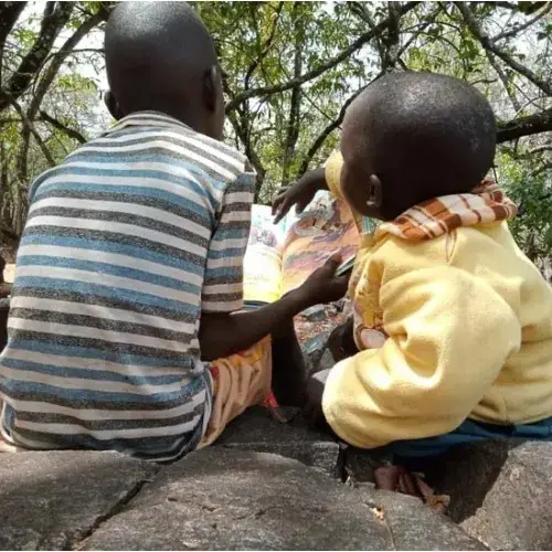 Two children seen from behind, sitting close together on a large rock beneath trees, looking at something in front of them