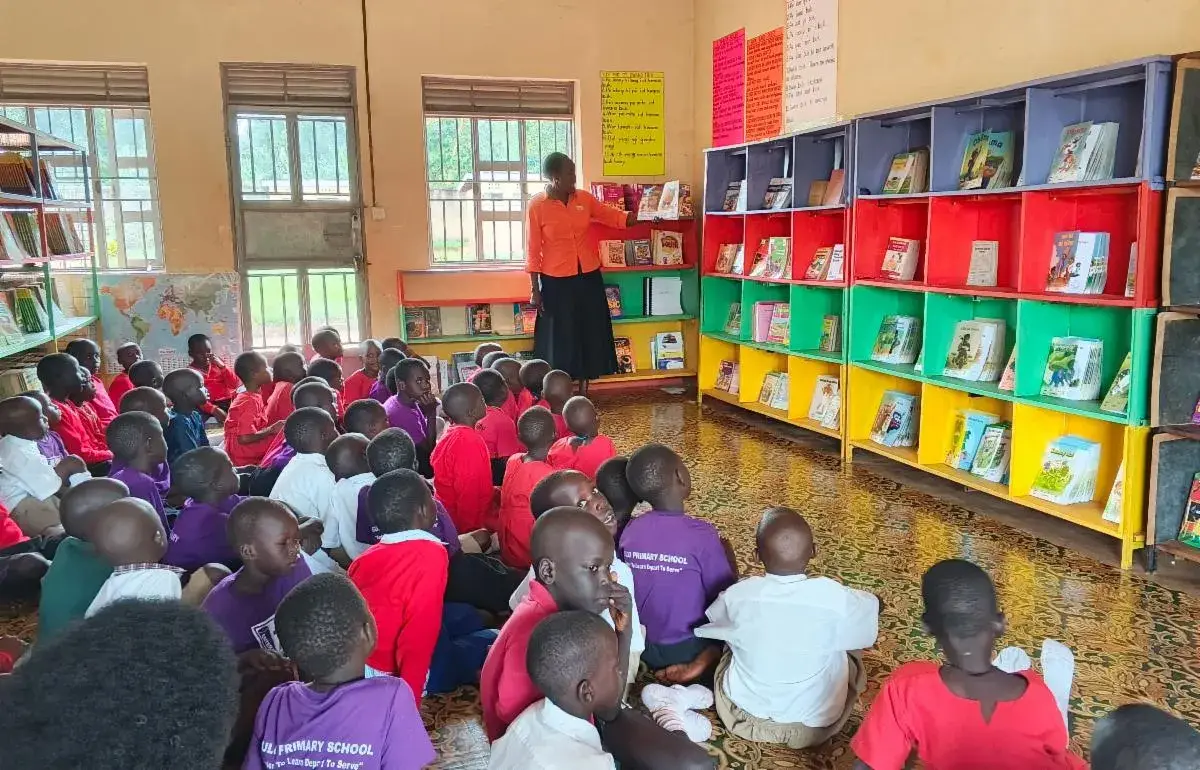 Pupils in school uniform sitting on a classroom floor in front of a wall of colourful book shelves, with a teacher standing at the back