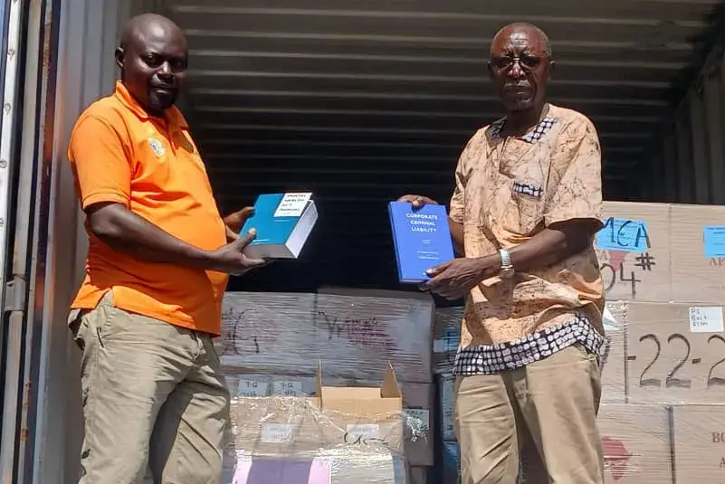 Two men in front of a shipping container holding up books, one in an orange shirt and one in a patterned shirt, with boxes stacked behind them