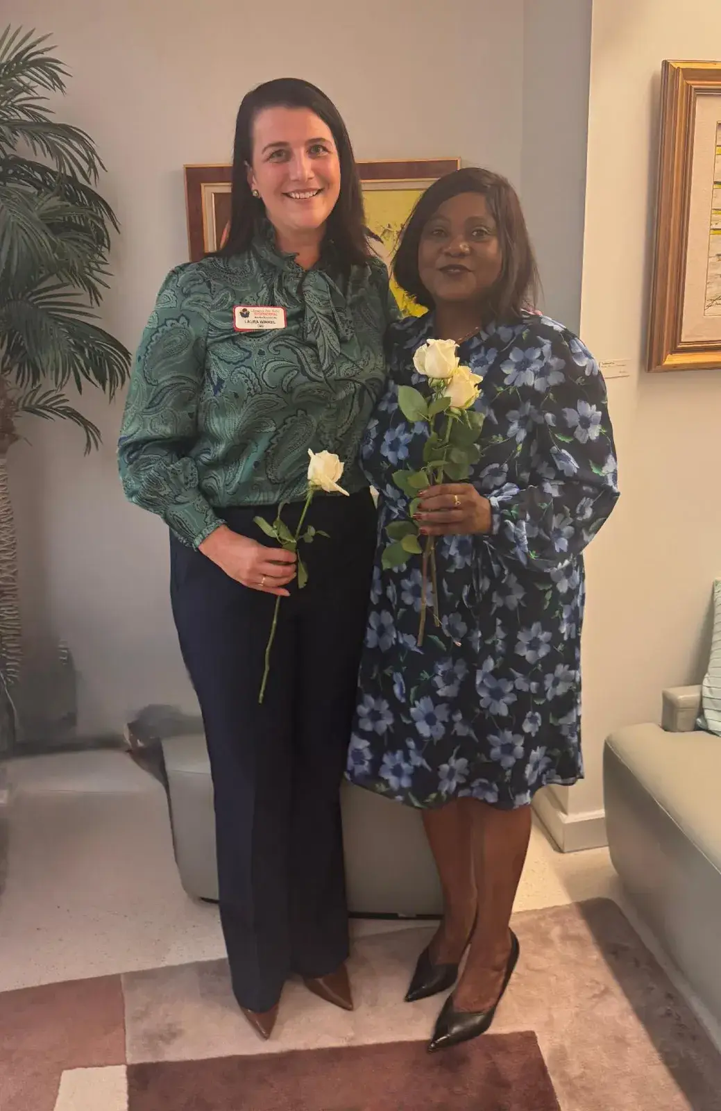 Two women standing side by side in an office, each holding a white rose: one in a green top and one in a patterned dress