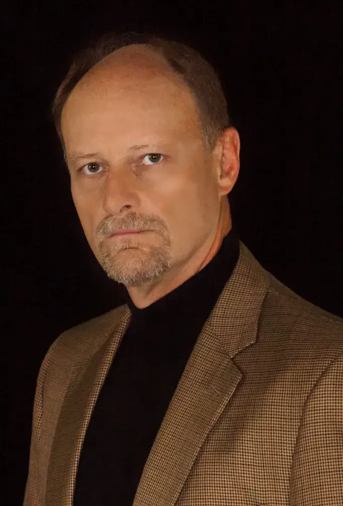 Brian Thiem, the author, in a brown jacket and black polo neck, in a portrait against a dark background