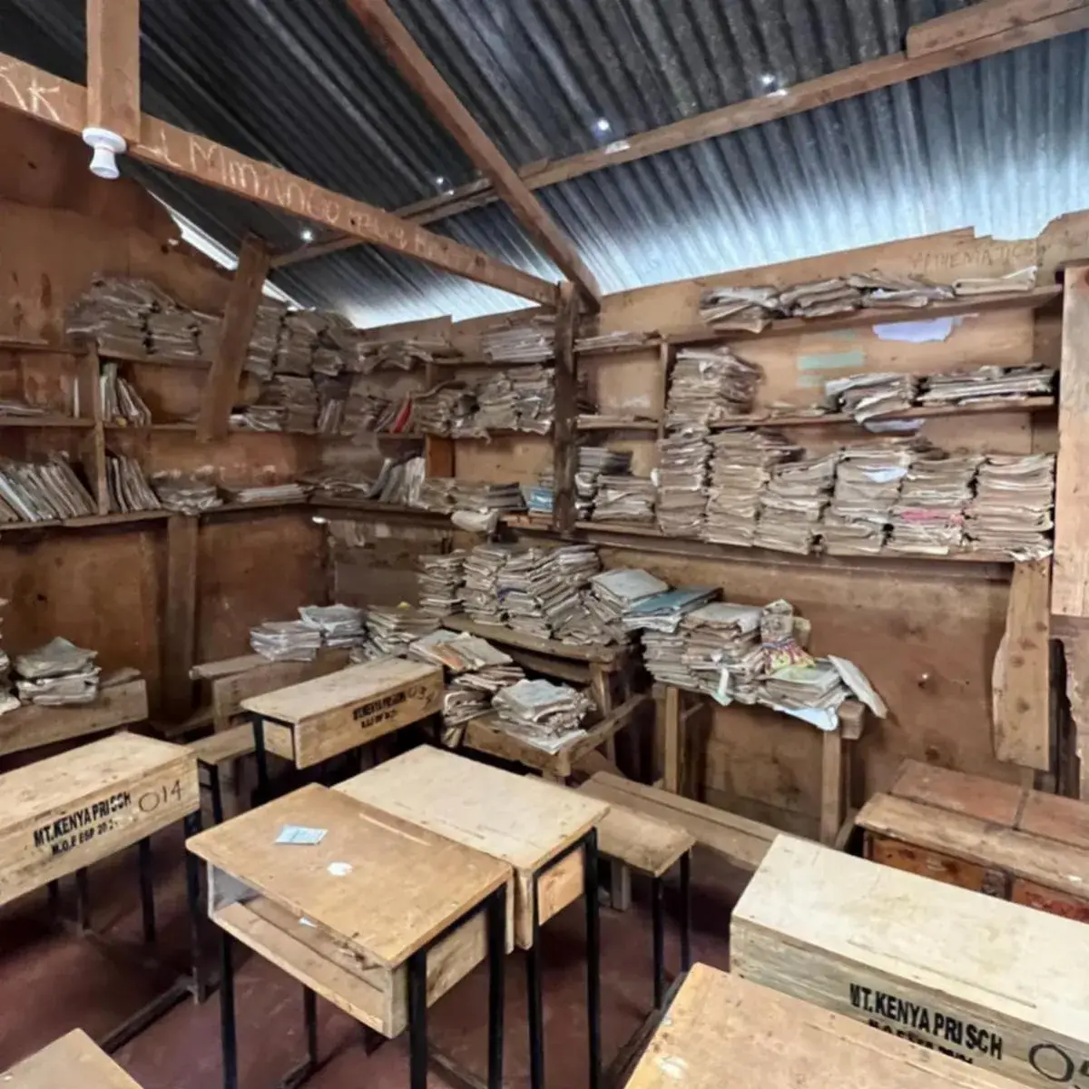 Inside a rural school library: rows of wooden desks and benches under a tin roof, with shelves of books and papers along the walls