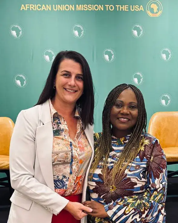 Two women standing close together in front of a green backdrop printed with the African Union Mission to the USA, one in a cream blazer and one in a patterned dress