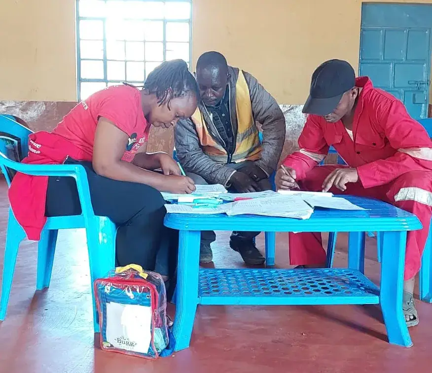 Four people around a blue table in a school yard: a woman in a red top writing on papers, a man in a dark jacket standing behind her, and a man in red overalls and a cap, with a child's school bag on the table