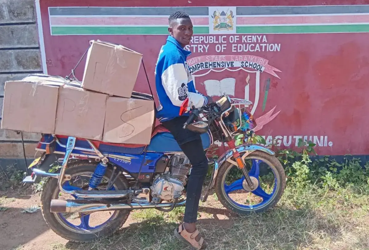 A man in a blue jacket on a motorbike loaded with cardboard boxes, in front of a red wall with the Kenya Ministry of Education crest