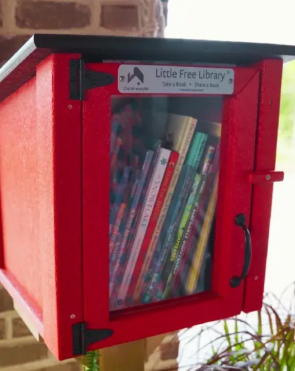 A red Little Free Library box with a glass door, filled with books, mounted on a post