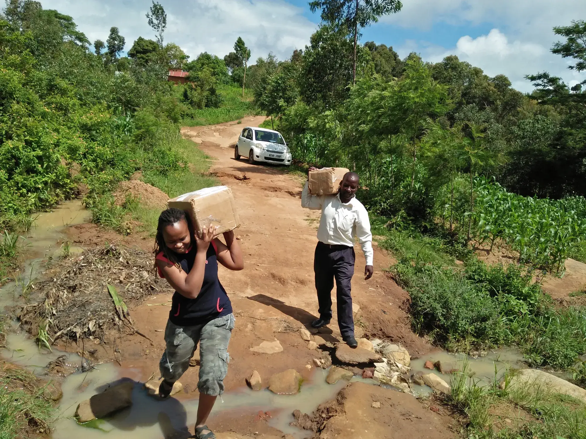 Wanjiku Francis, the Kenya Operations Coordinator, and a teacher carrying boxes of books across a muddy stream on a dirt road, a car parked on the road behind them