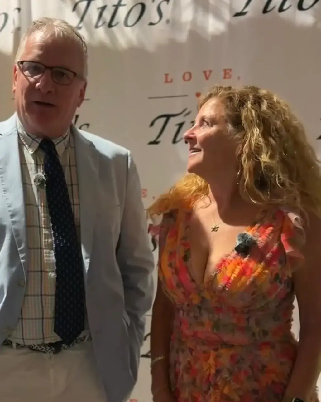A man in glasses and a light blue jacket beside a woman with long, curly blonde hair, who looks up at him
